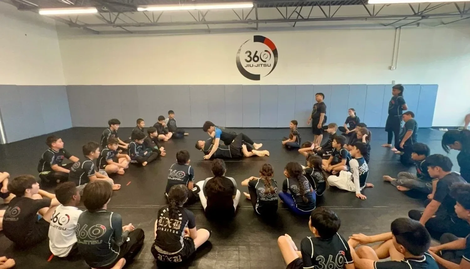 A full youth class circled up on the mats under the 360 Jiu-Jitsu wall logo